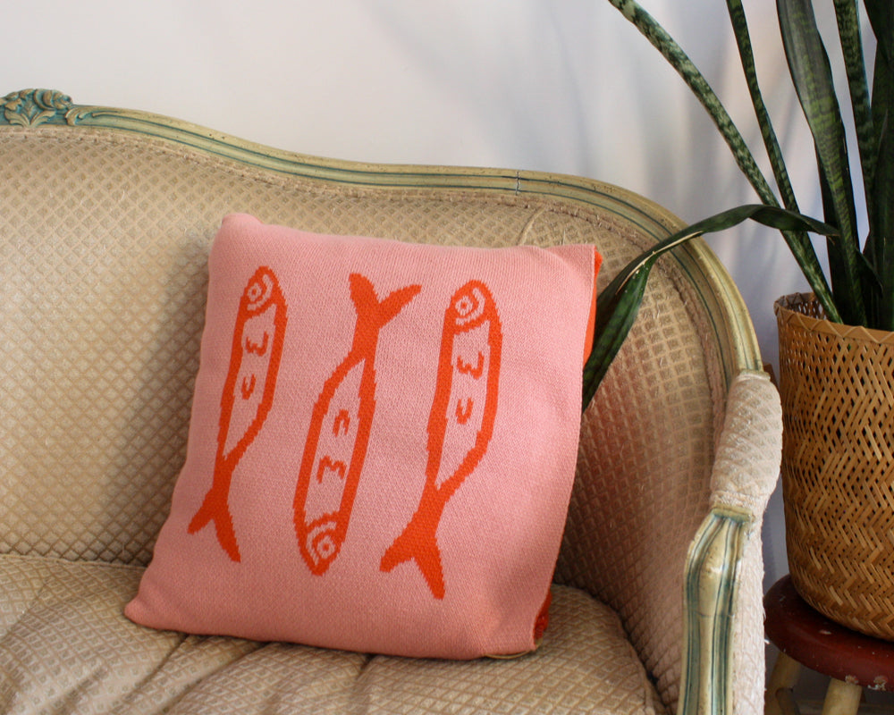 A cozy, square throw pillow featuring a pink background with three orange fish motifs arranged vertically. The design is playful and modern, adding a touch of whimsy to the textured beige sofa it's resting on. Perfect for a unique, artistic home decor touch.