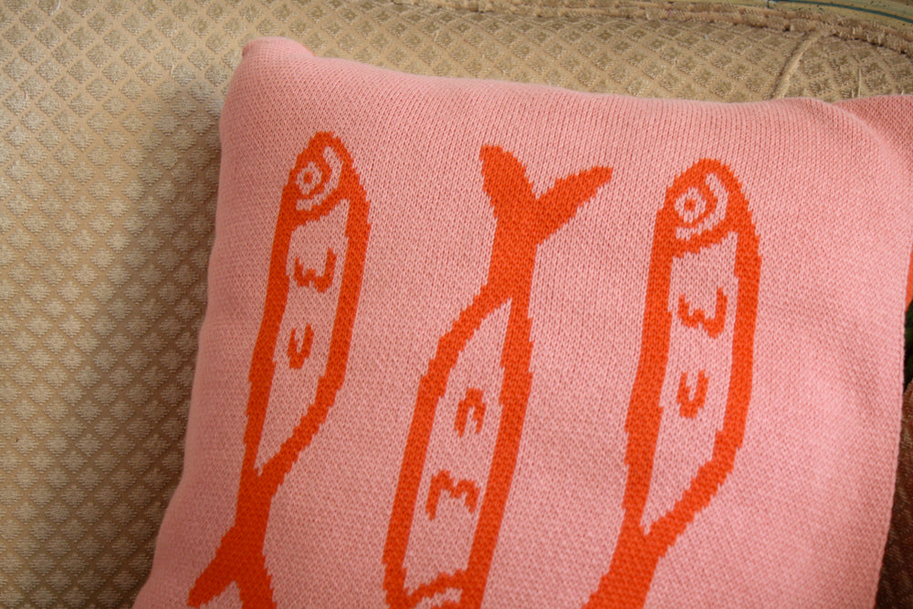 A cozy, square throw pillow featuring a pink background with three orange fish motifs arranged vertically. The design is playful and modern, adding a touch of whimsy to the textured beige sofa it's resting on. Perfect for a unique, artistic home decor touch.